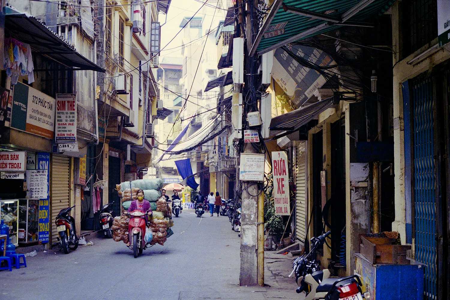 35mm photo of the streets of Hanoi with a lady on a scooter transporting a lot of bags in vietnam